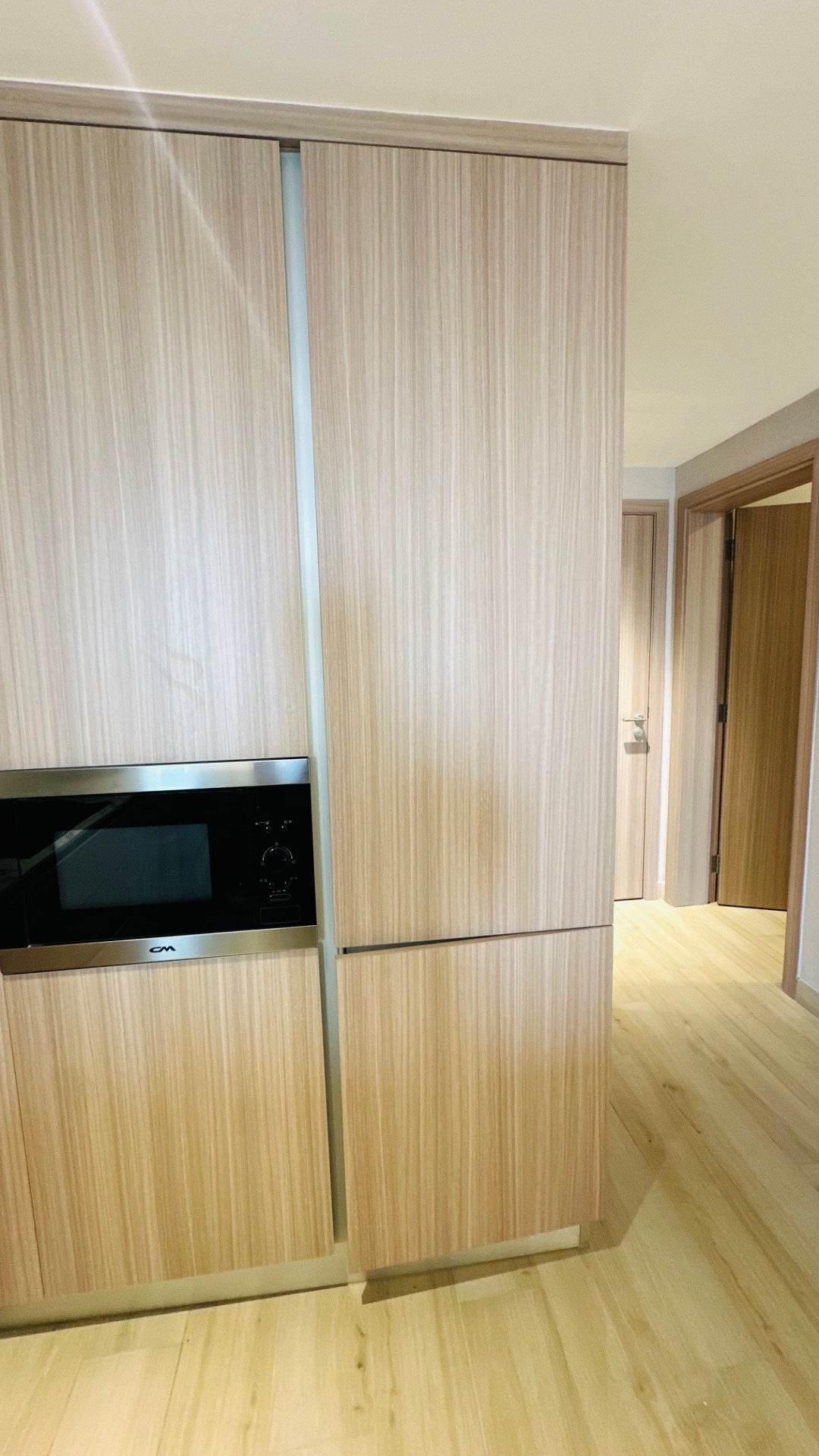 Light wood veneer cabinetry with an integrated stainless steel microwave, opening onto a hallway with matching laminate flooring and flush doors