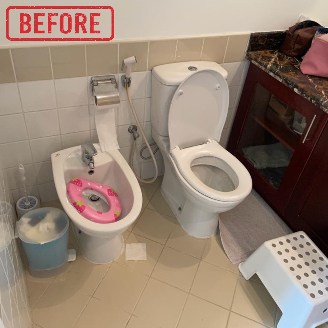 A cluttered and dated compact bathroom features a white toilet and bidet with a child's seat, a dark marble-topped vanity, and light ceramic