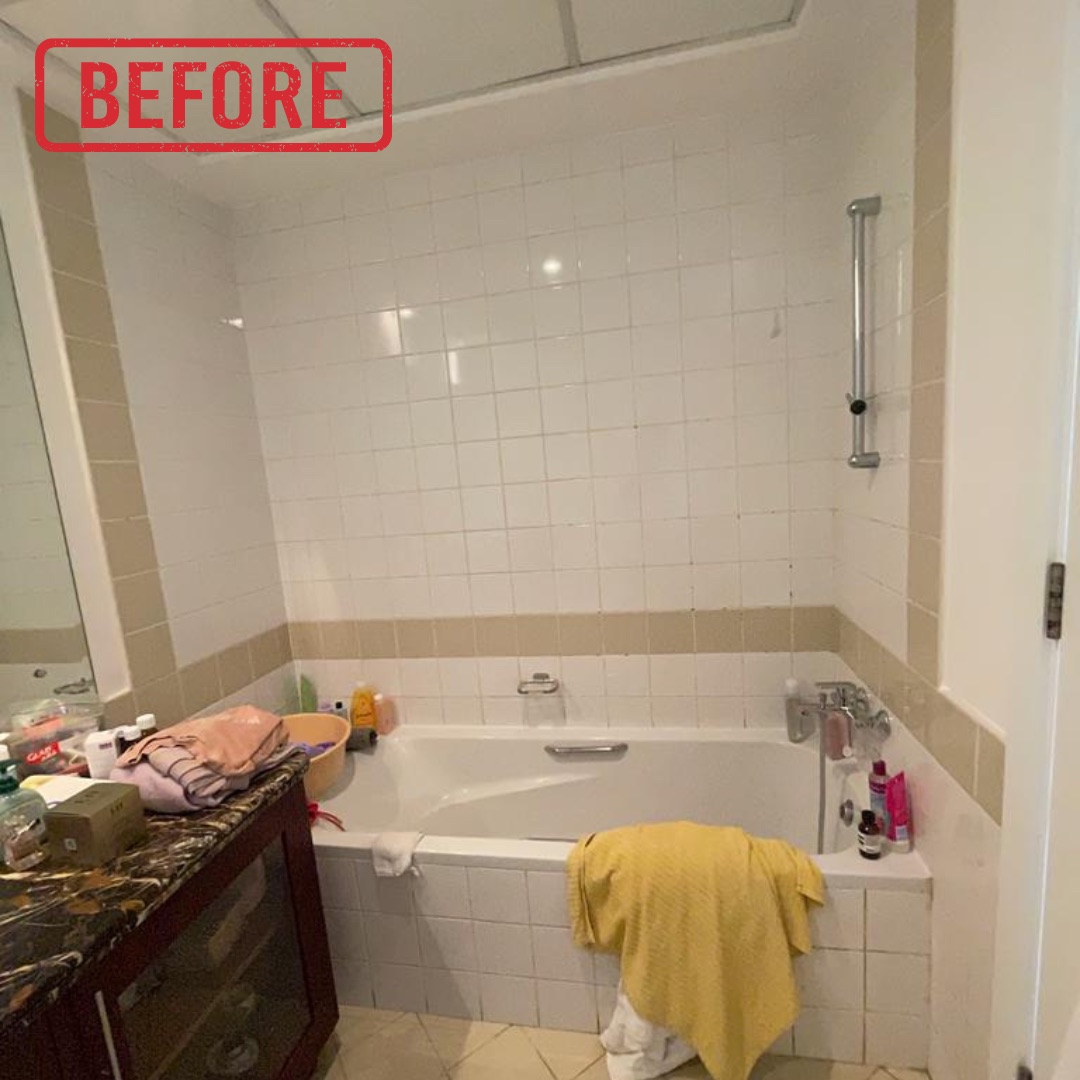 A wide-angle "before" shot of a cluttered and dated bathroom featuring a built-in bathtub, two-tone tiled walls, a dark marble-topped vanity