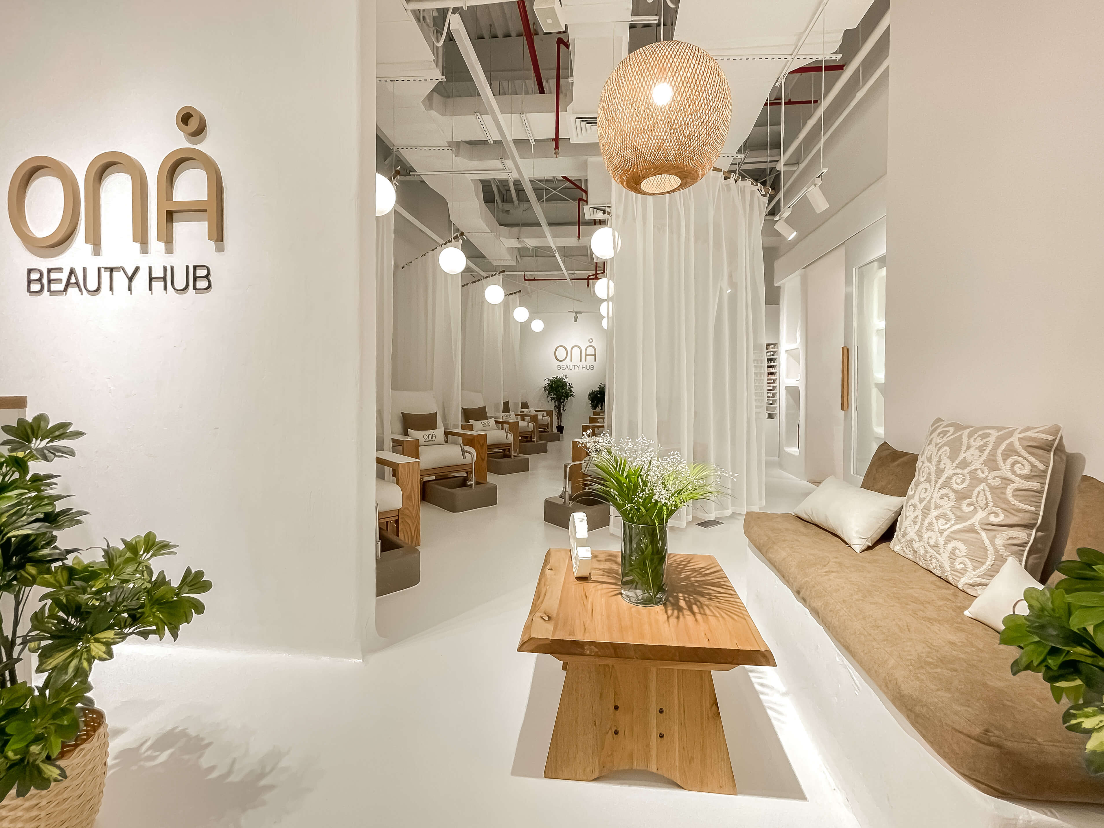 Pedicure stations with light wood chairs and privacy curtains under an exposed ceiling in a beauty hub interior
