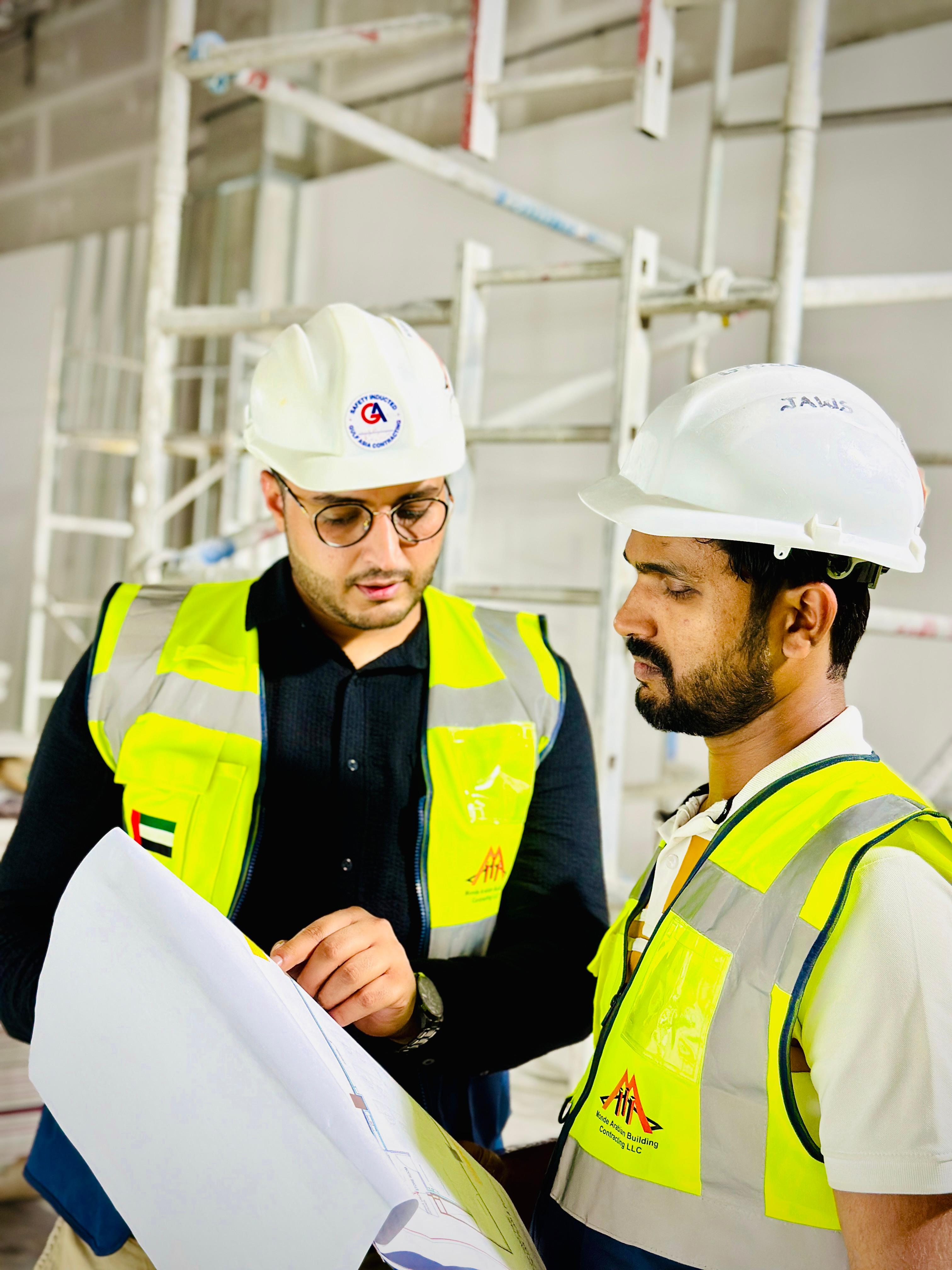Two construction project managers or engineers, wearing hard hats and high-visibility vests, reviewing blueprints