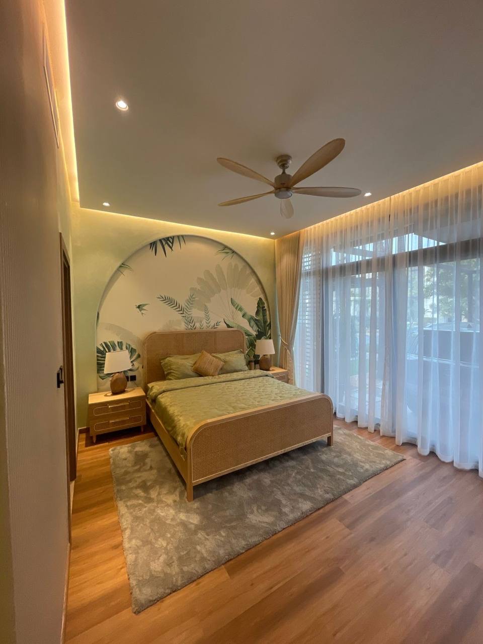 A bright and serene modern bedroom featuring a rattan bed frame with green bedding, wood flooring, a tropical-themed wall mural