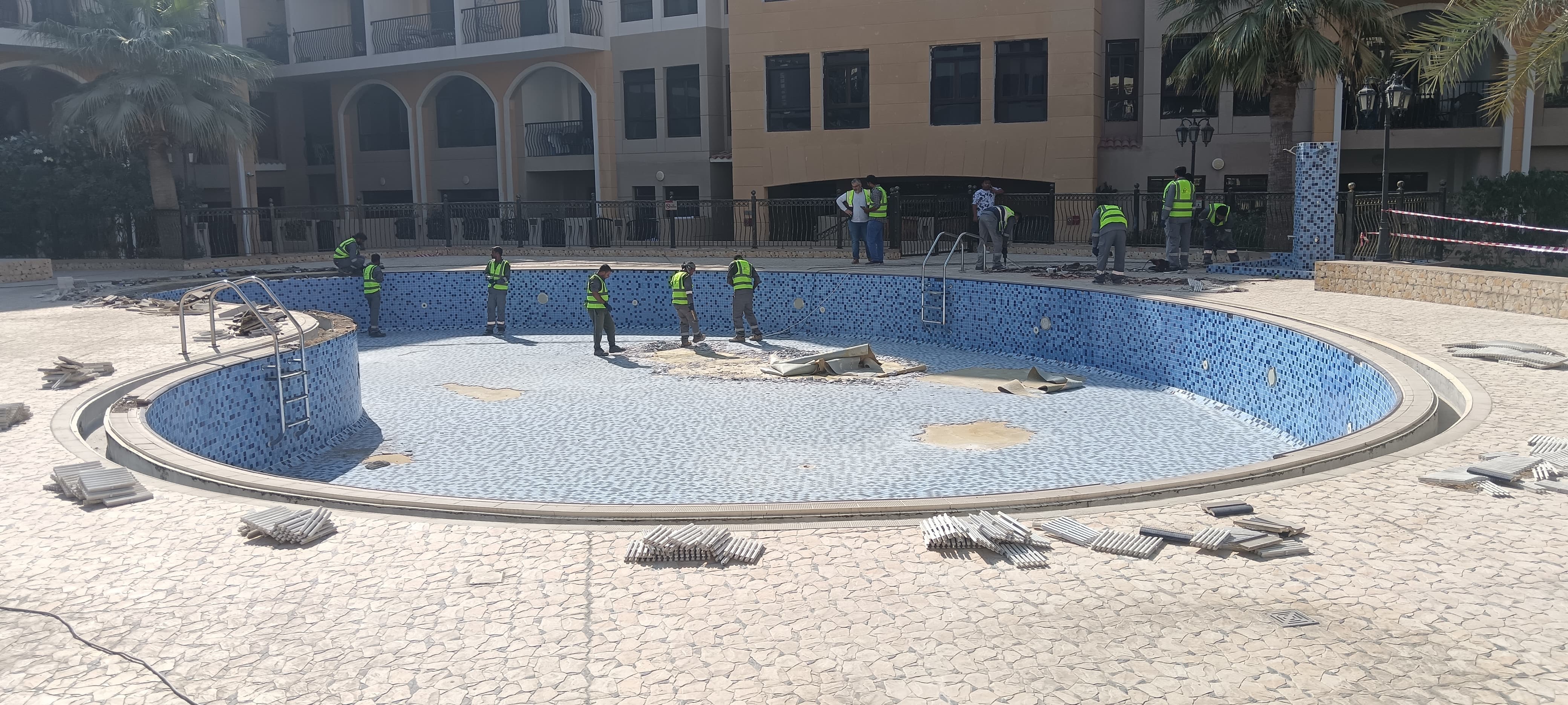 Team of workers renovating a large swimming pool shell with blue mosaic tiles