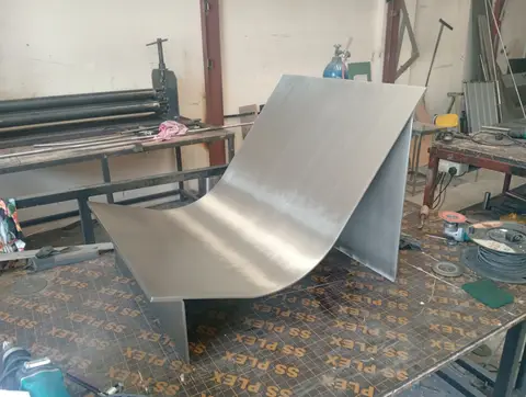 A large, expertly curved brushed metal sheet, likely a component for a modern chair or bench — APEX STEEL WORKS L.L.C