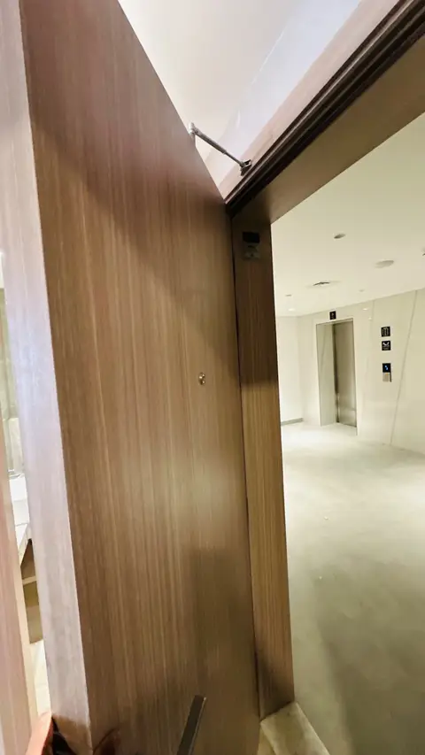 Modern light-wood door opening into a white-walled hallway with elevator lobby