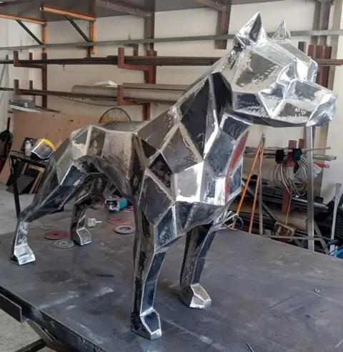 A striking geometric metal sculpture of a dog, likely a pitbull, stands prominently on a workbench — APEX STEEL WORKS L.L.C