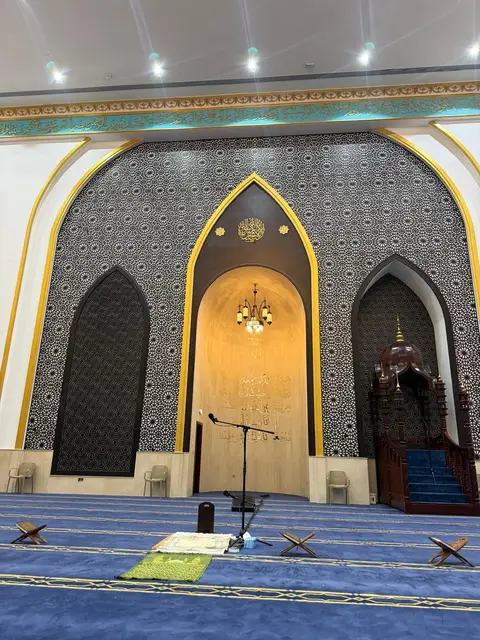 A grand mosque prayer hall features a central mihrab with Arabic calligraphy, a warm chandelier, patterned — Malak Afzal