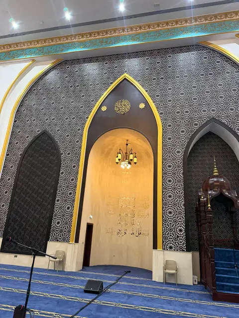 An ornate mihrab with golden Arabic calligraphy, intricate dark geometric wall patterns, a traditional wooden — Malak Afzal