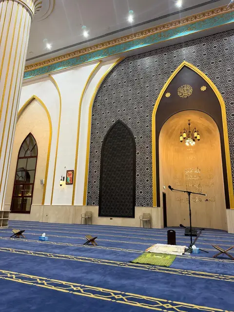 A deep blue carpet with gold patterns leads to an ornate mihrab with a warm chandelier and Arabic calligraphy — Malak Afzal