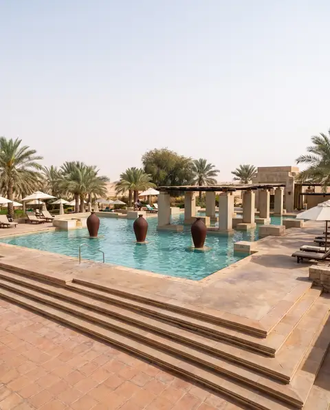 An expansive outdoor swimming pool at a luxurious desert resort features traditional terracotta-style urns, stone pillar