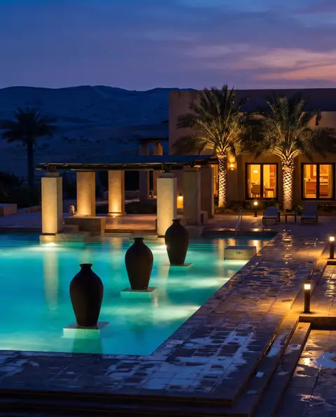 A luxurious desert resort pool glows with warm artificial light at twilight, set against a serene backdrop of rolling sa