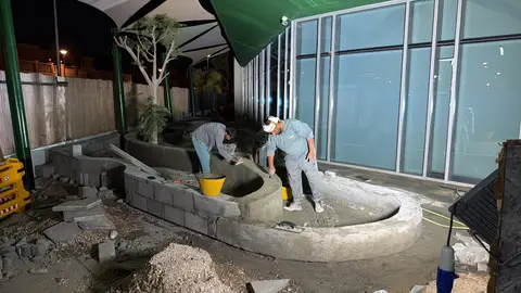 Three construction workers diligently apply cement to a newly formed, curved concrete planter structure at night, set ag