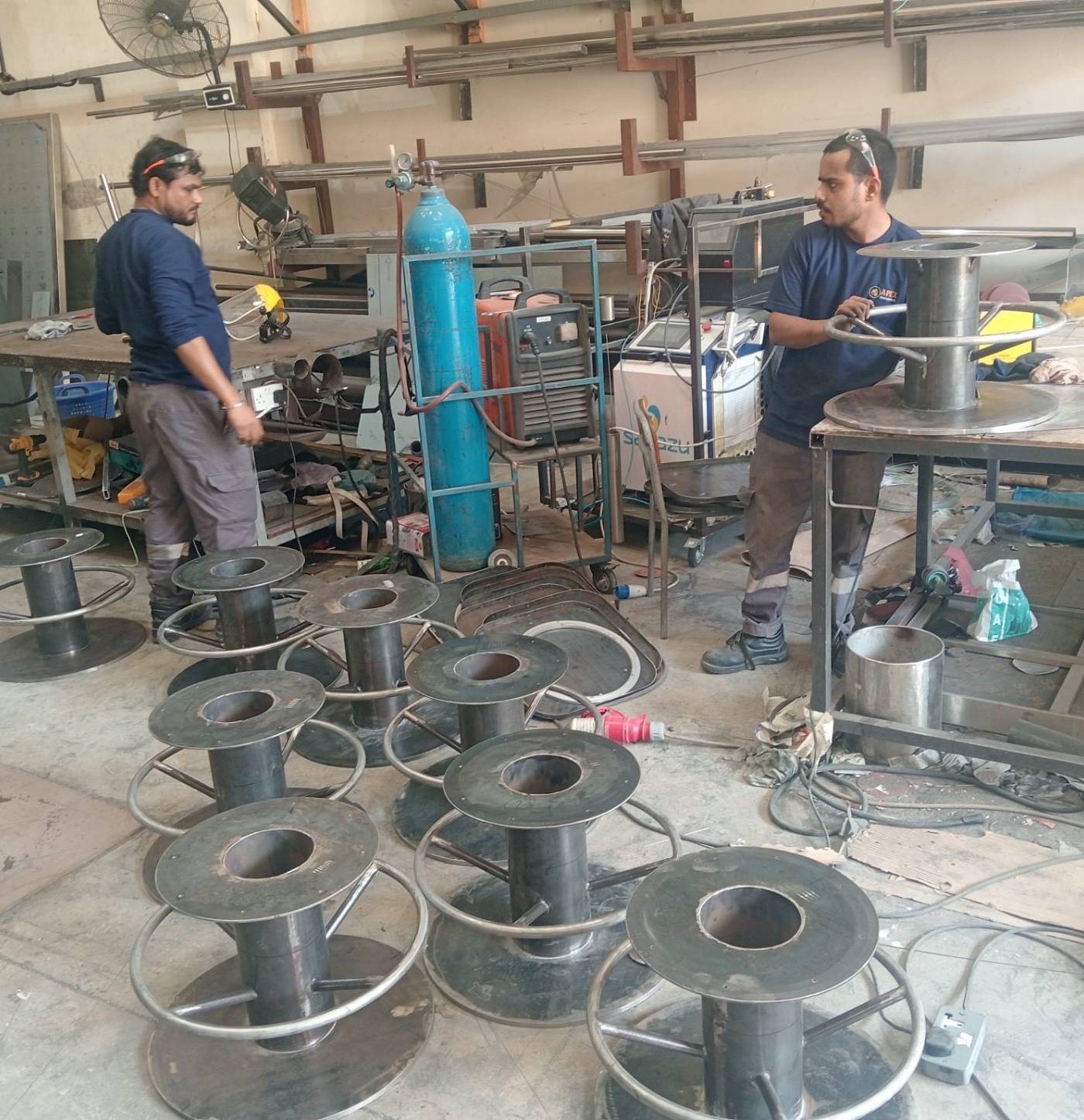 Two fabricators assembling steel stool bases among welding equipment and raw material in an industrial workshop