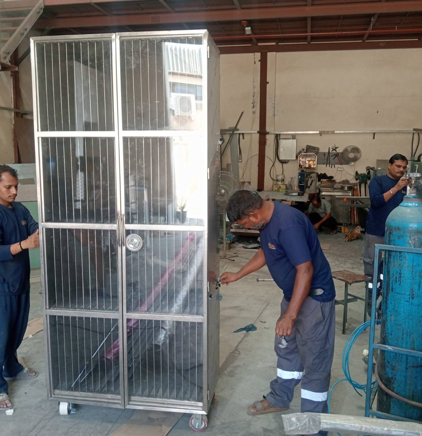 Workers fabricating a tall stainless steel cabinet with mesh doors and heavy-duty castors in a busy industrial workshop