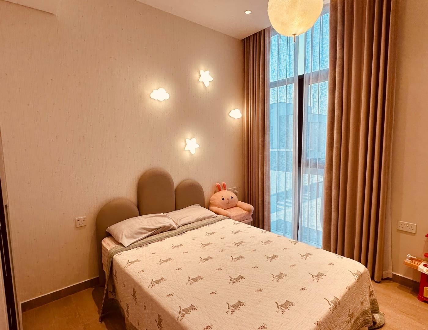 Arched upholstered headboard with cloud and star wall lights, a large window with beige curtains and a plush bunny toy resting on a stool beside the bed