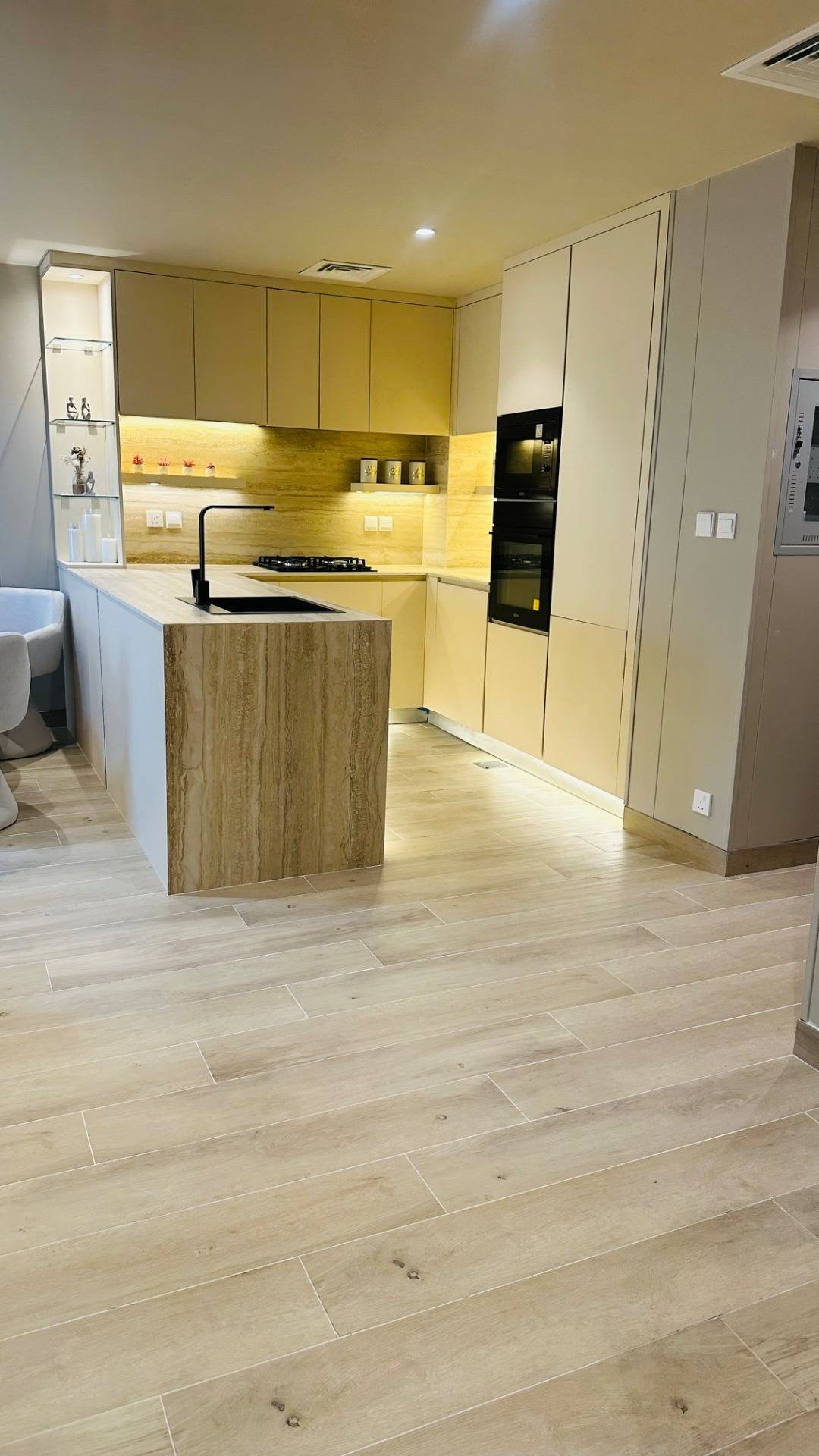 Freshly renovated contemporary kitchen with light beige cabinetry, stone clad island with an integrated black sink and warm under cabinet lighting on a matching stone backsplash