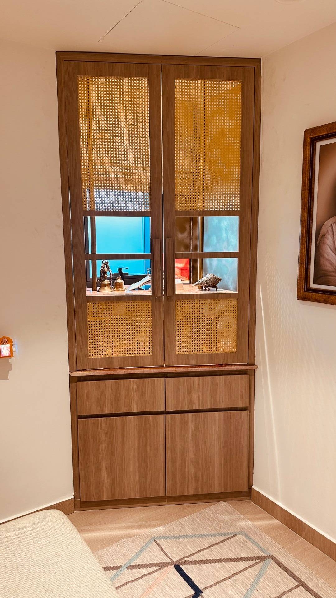 Wooden cabinet with cane webbing inserts on glass fronted doors, brass decor items displayed on top and solid wood drawers below, against a warm white wall with a patterned rug.