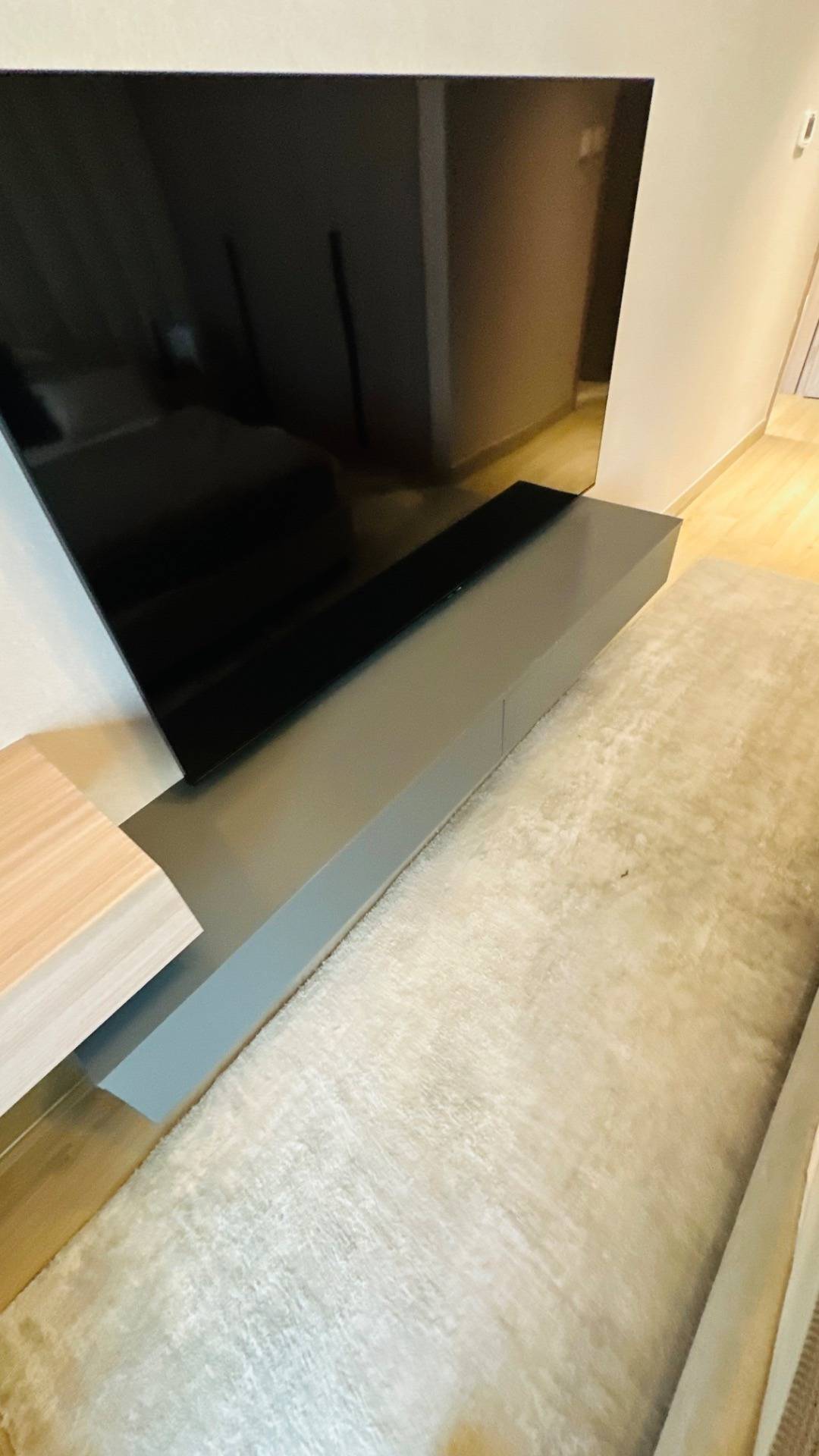 Modern media wall with a wall mounted television above a grey floating console, a light wood accent shelf and a beige shaggy rug on light wood flooring