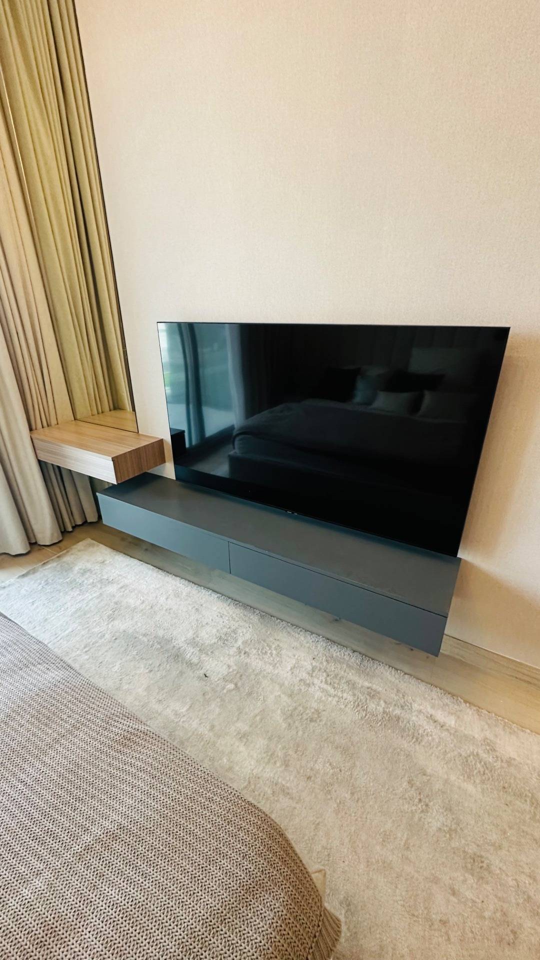 Dark grey floating media console below a wall mounted television with a light wood floating shelf, beige curtains and a textured rug in a modern bedroom.