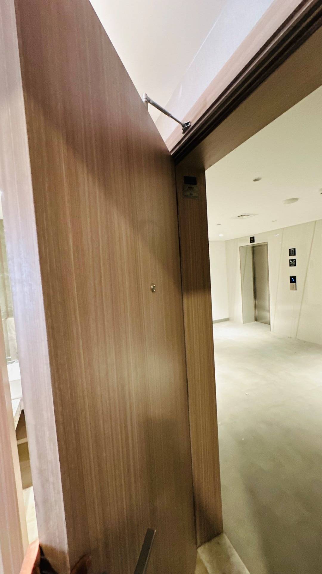 Light wood grained door with a sleek door closer opening into a white hallway with a polished stainless steel elevator and recessed ceiling lights
