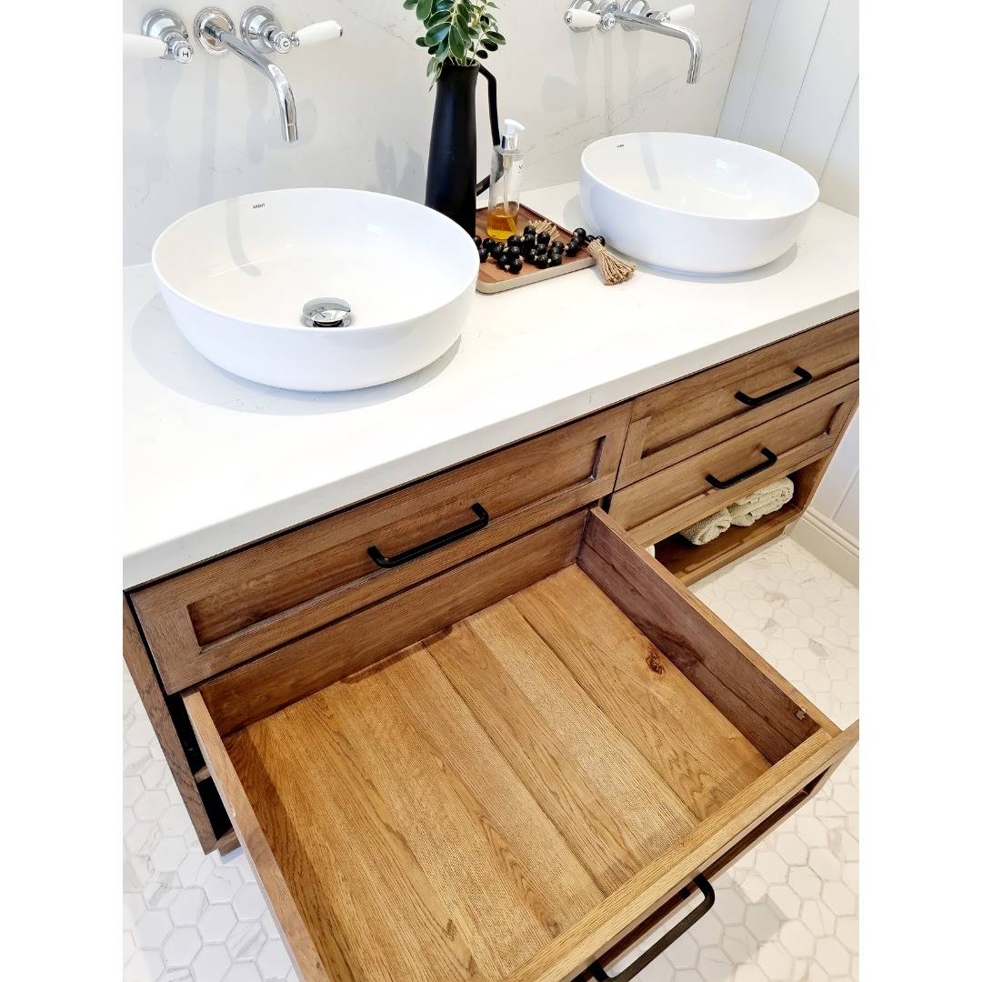 Bright renovated bathroom with double vanity, white vessel sinks, wall-mounted chrome faucets, white marble countertop atop light wood cabinetry, white hexagonal floor tiles, and open drawer showing natural wood interior