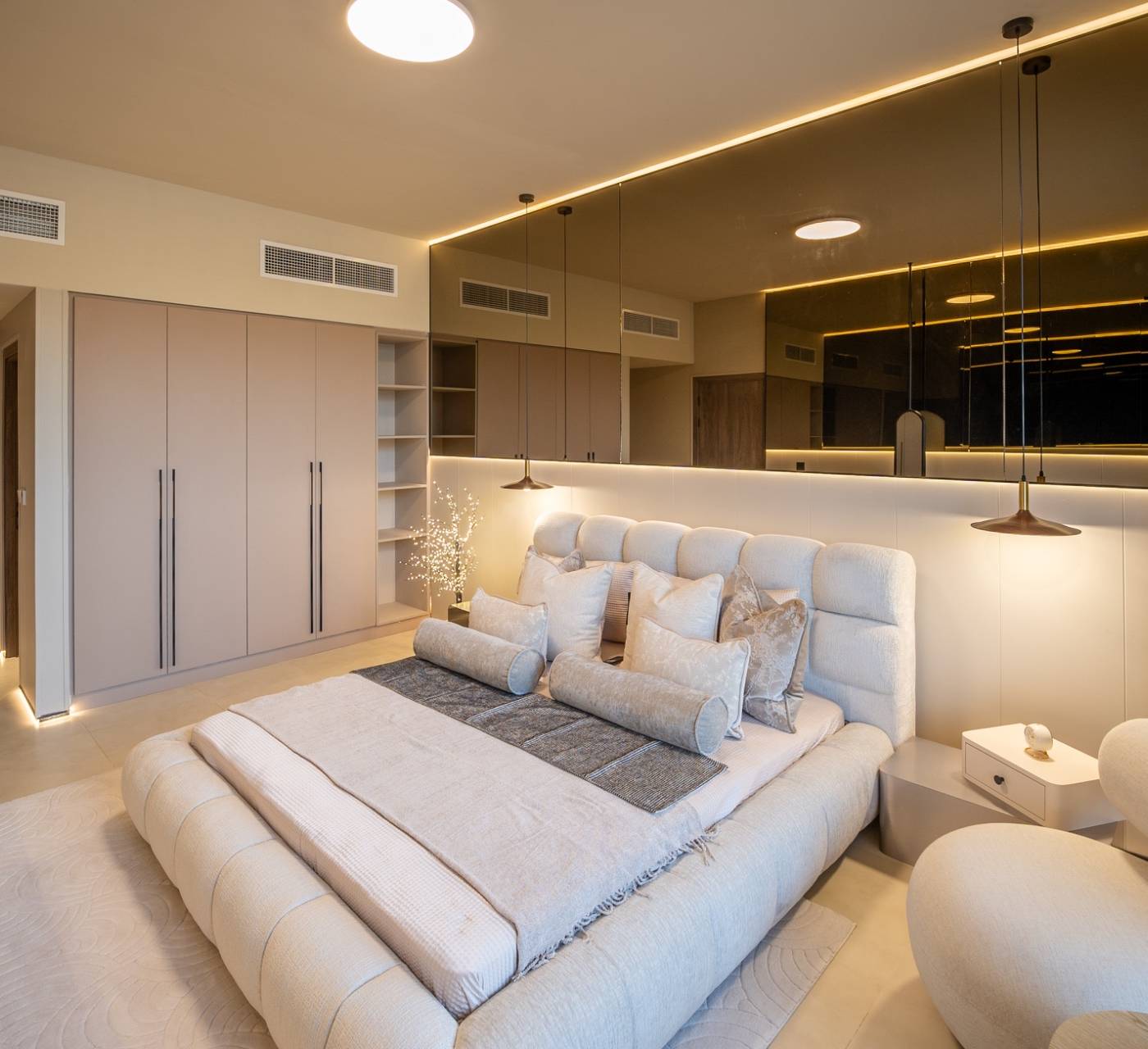 Luxurious modern bedroom with plush upholstered bed, pendant lights, mirrored accent wall with strip lighting, and built-in wardrobes — villa remodeling Dubai master suite.