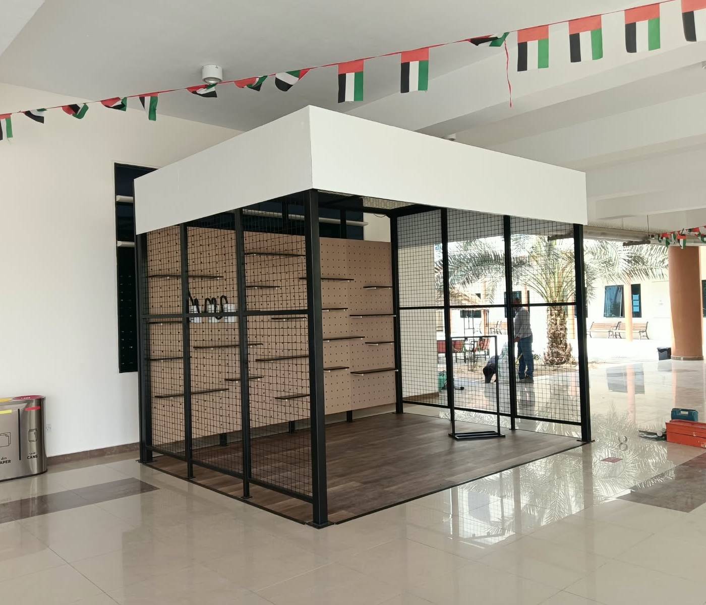 Modular display kiosk with black metal grid framing and pegboard shelving, plus stainless steel waste segregation bins, fabricated for a UAE fit-out