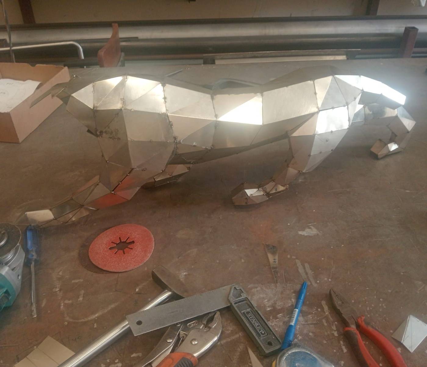 Faceted stainless steel sculpture of a big cat with visible weld marks on a cluttered workshop bench at APEX STEEL WORKS, surrounded by an angle grinder, pliers and a hammer.