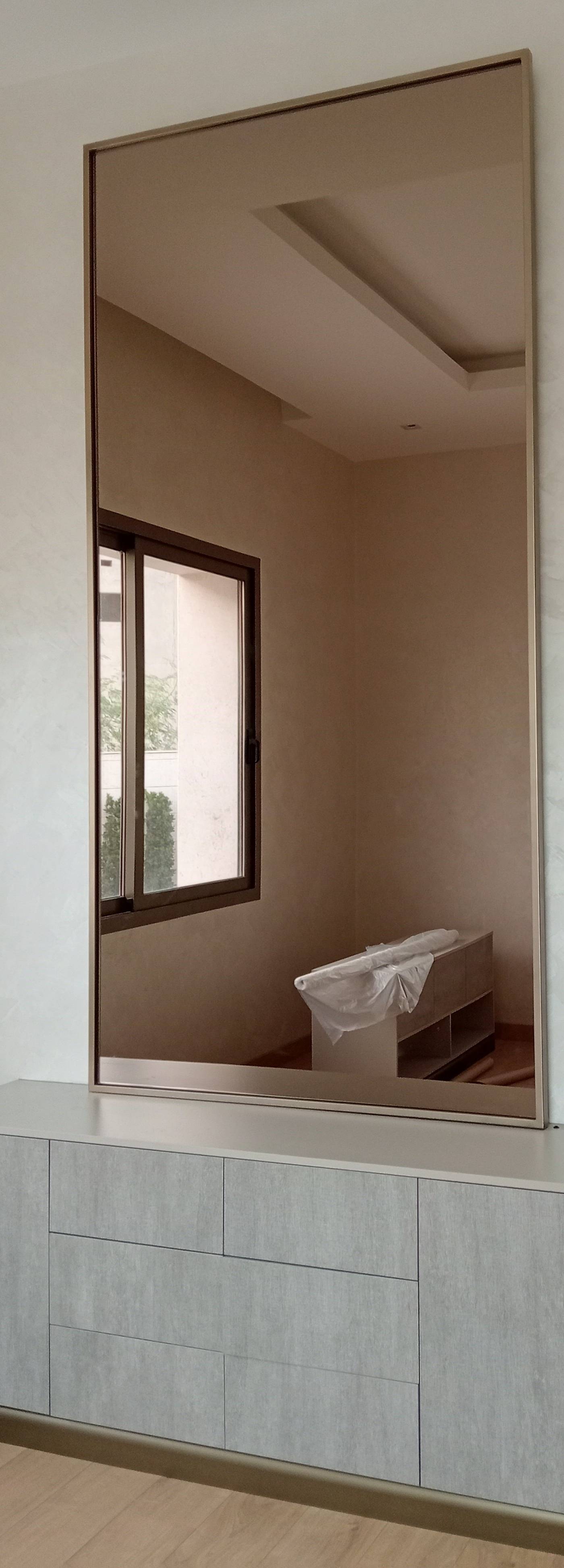 A full length mirror with a bronze frame stands on a light grey integrated storage cabinet against a textured off white wall and warm wood flooring