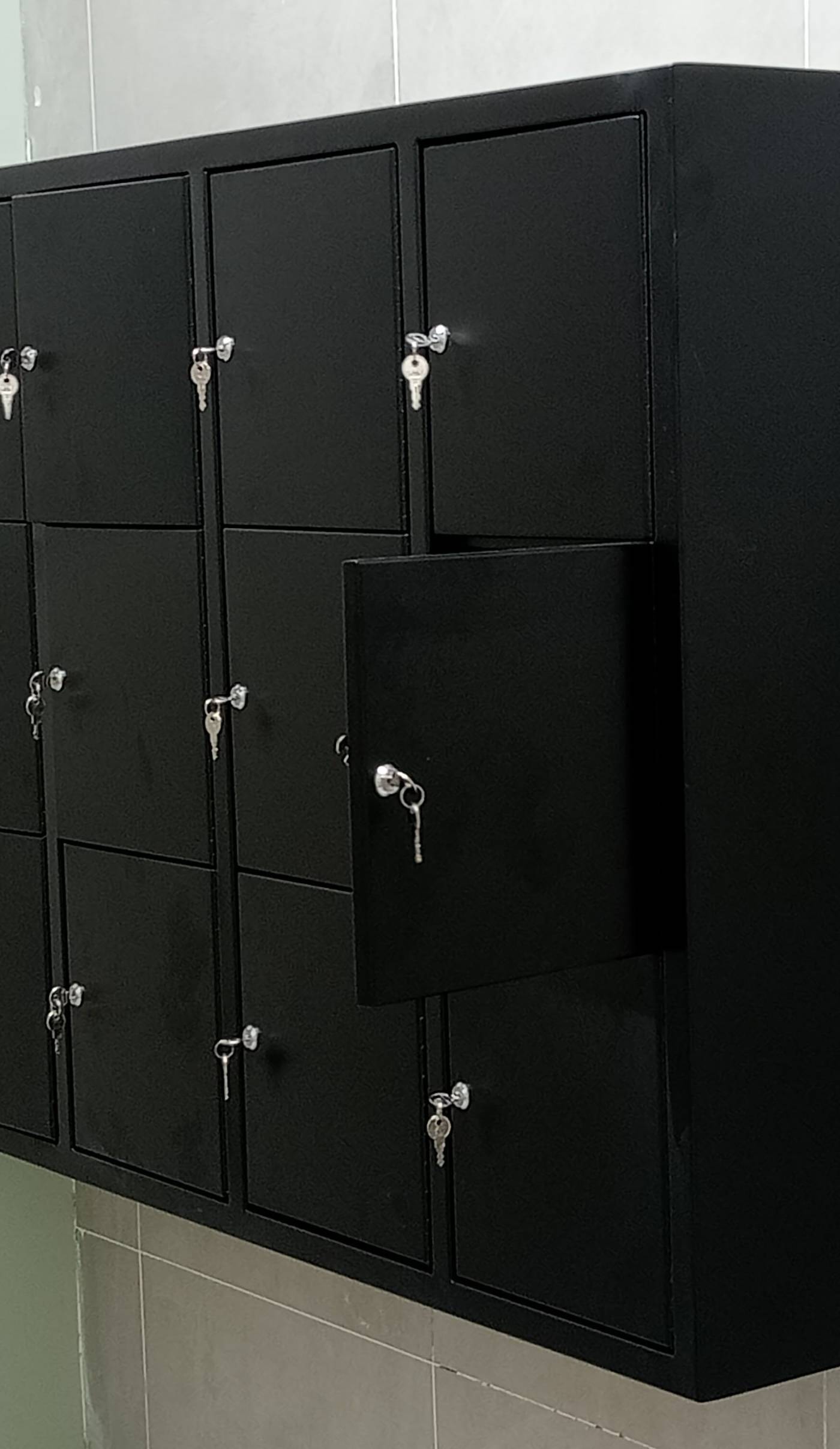 A bank of sixteen wall-mounted black metal lockers with keys in the locks against a light grey tiled wall