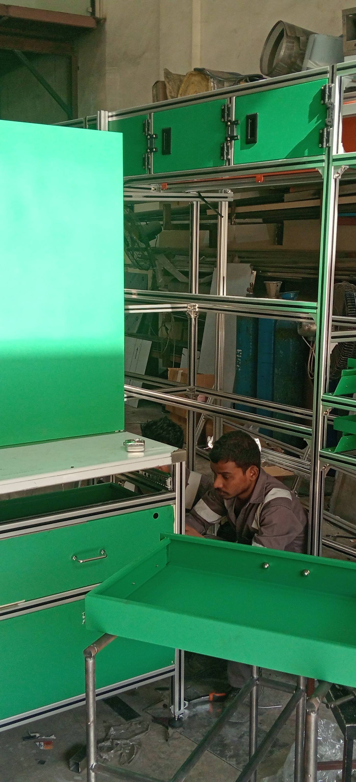 A busy industrial workshop shows a worker assembling green and silver modular structures during custom shelving and workstation fabrication