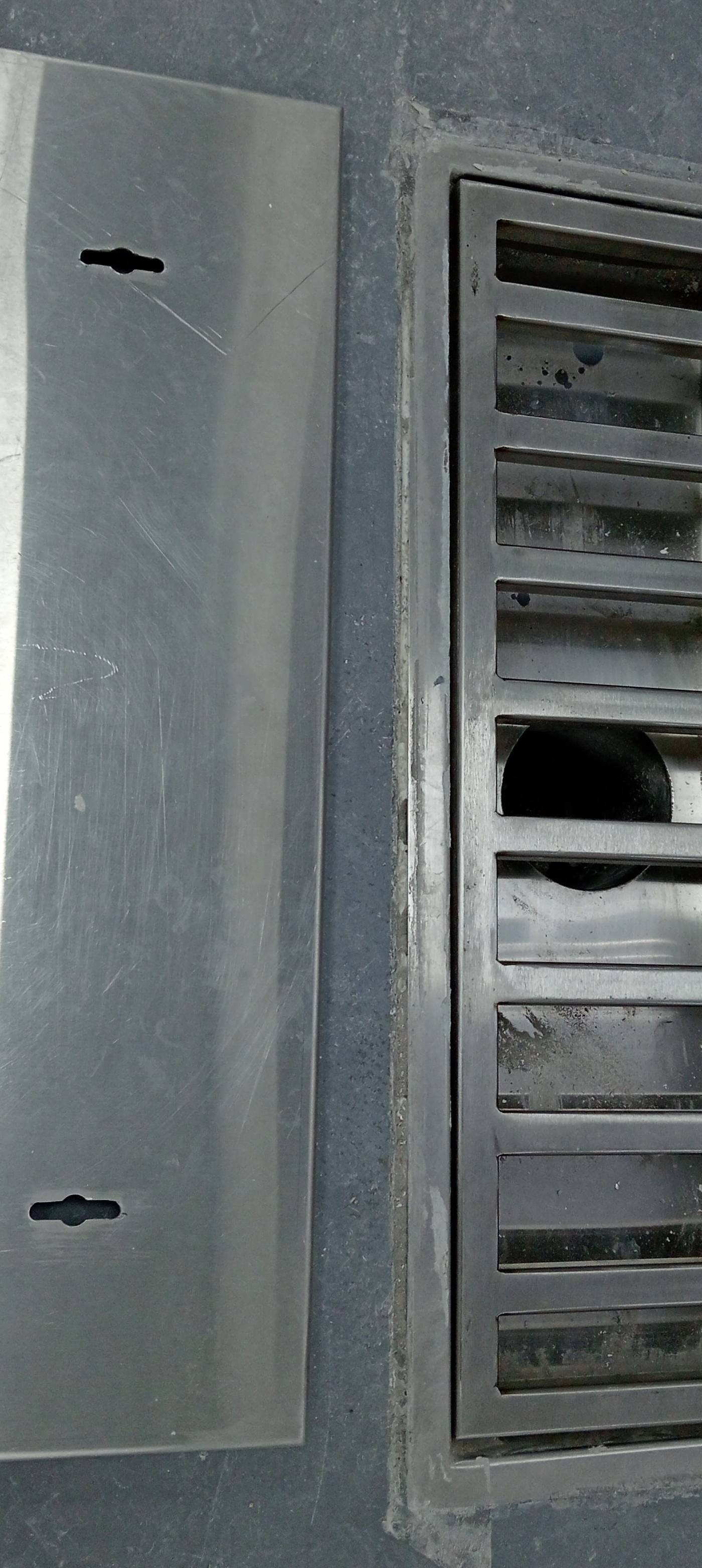 Stainless steel linear floor drain with its cover plate resting beside it on a dark grey concrete floor