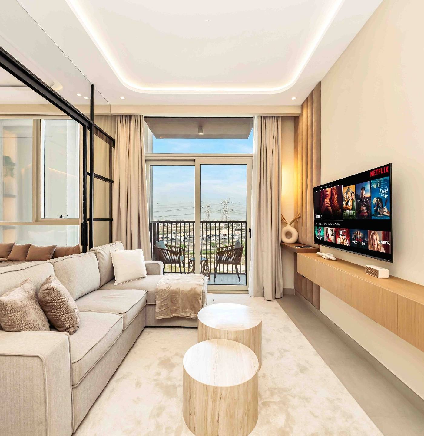 Modern Dubai living room with an L-shaped sofa, two round light wood coffee tables on a plush rug and a wall mounted TV above a floating media console.