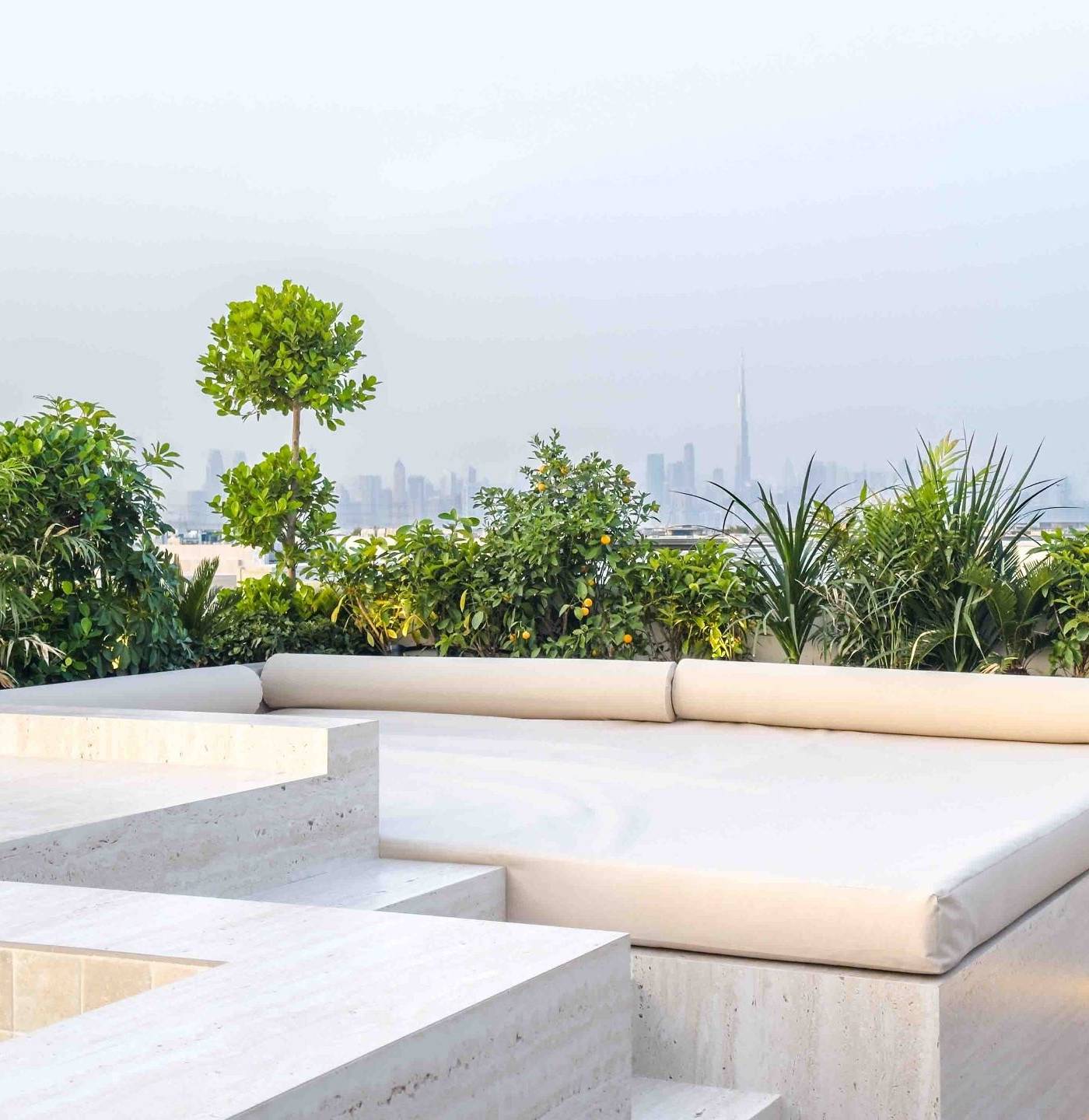 Rooftop terrace with a built-in travertine daybed, stepped stone levels, green planting and the distant Dubai skyline including Burj Khalifa.