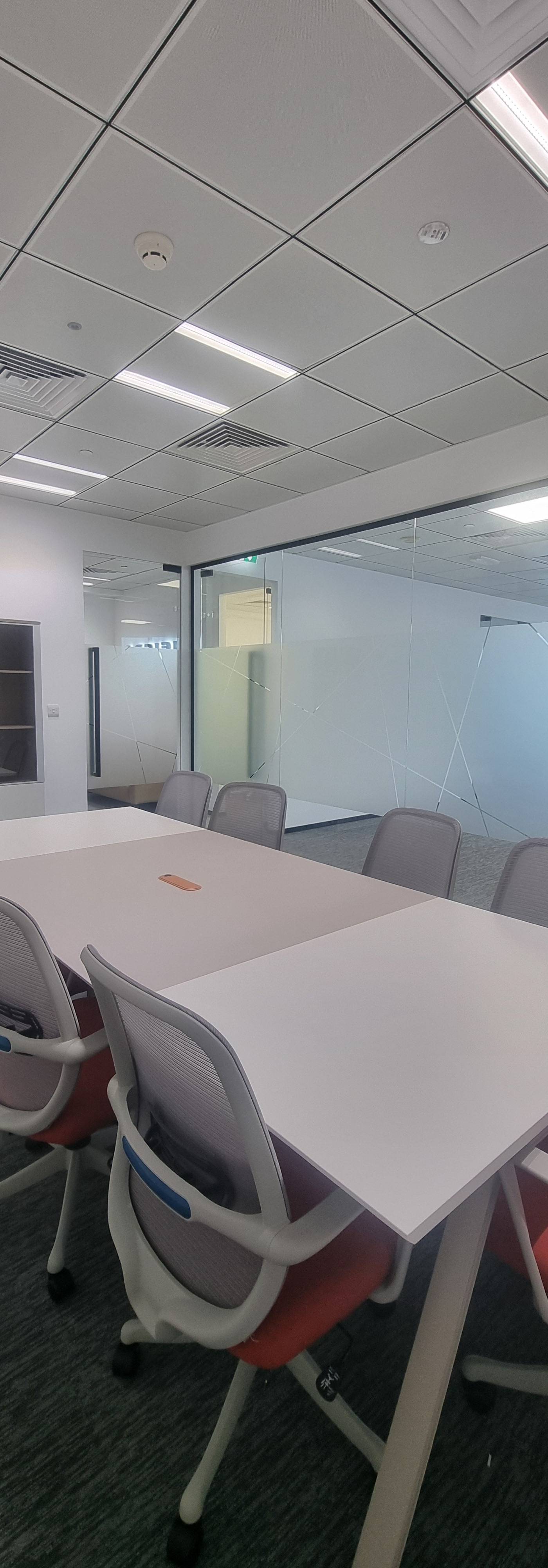 Meeting room with a long white table, orange ergonomic chairs, frosted glass partitions and white cabinets under a grid ceiling