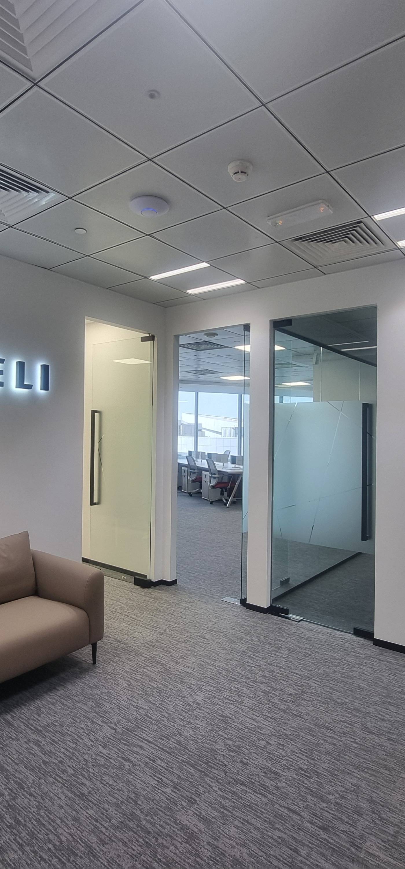 Office reception area with a backlit Aspireli logo on a white wall, beige seating and grey patterned carpet behind glass partitions