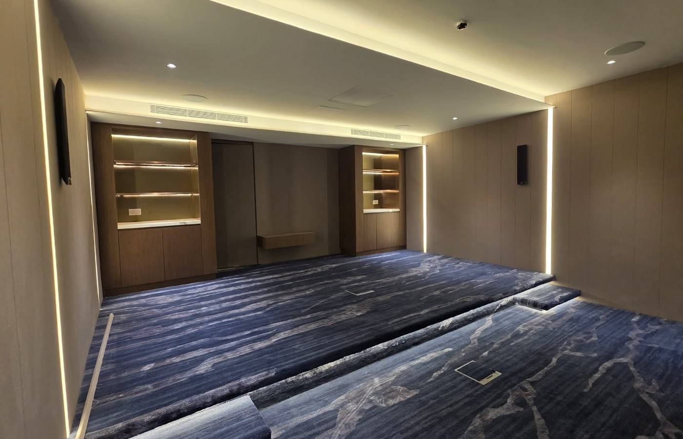 Built-in wooden cabinets with illuminated shelving and vertical LED strip lighting on textured beige wall panels, above a blue patterned carpet with a raised platform