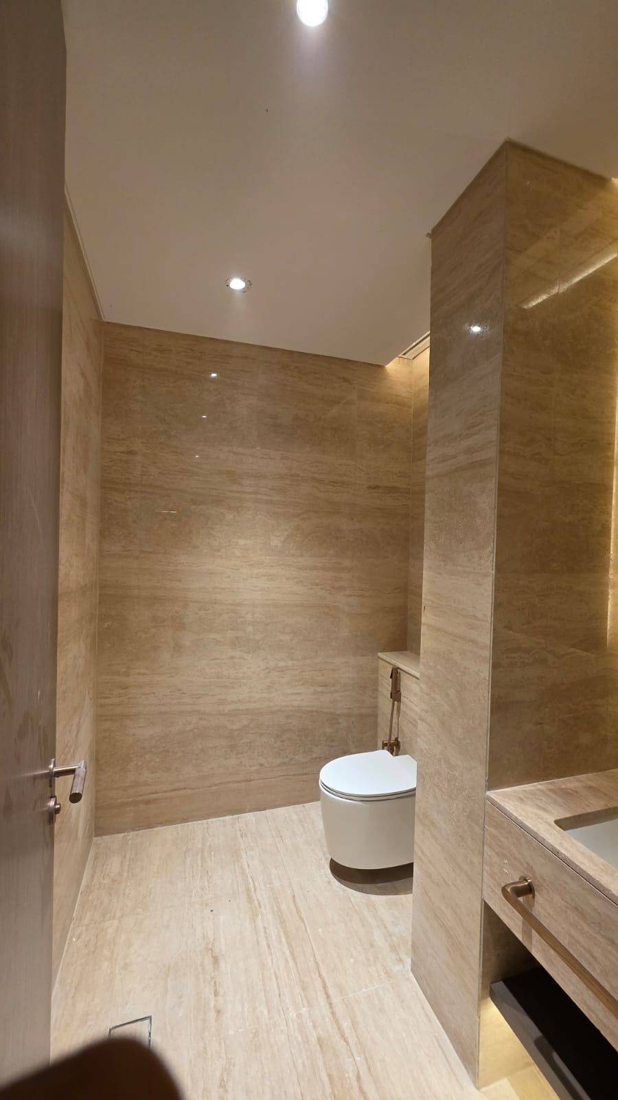 Commercial bathroom fit-out with warm beige veined marble walls and floors, a wall mounted toilet, bronze fixtures and a contemporary vanity under recessed and accent lighting