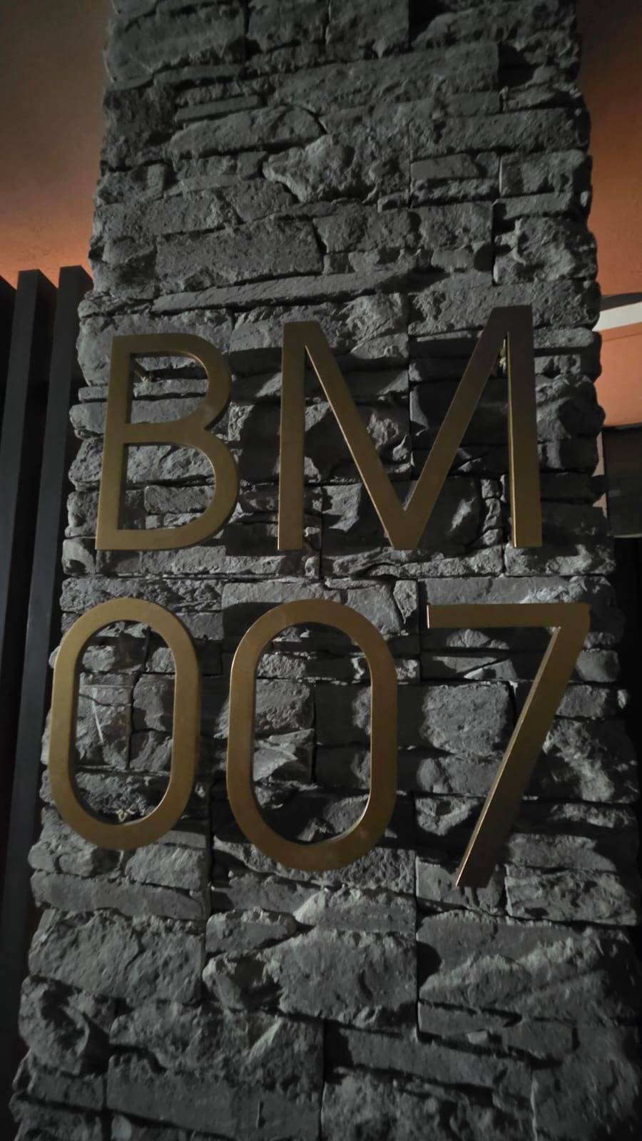 Close-up of a textured grey stacked stone feature wall with gold metallic lettering reading BM 007, lit by warm ambient light