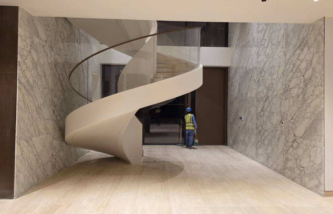 Grand spiral staircase with a glass railing in a marble-lined entrance hall with veined white walls and light stone flooring, photographed during final cleanup by the joinery team
