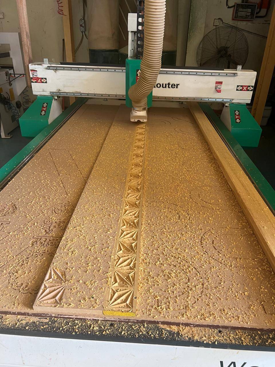CNC router carving intricate geometric patterns into a large timber panel inside a joinery workshop with sawdust in the air