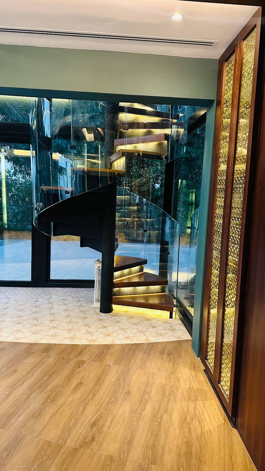 Modern spiral staircase with illuminated wooden treads and a clear glass railing beside full height windows and a custom dark wood feature wall