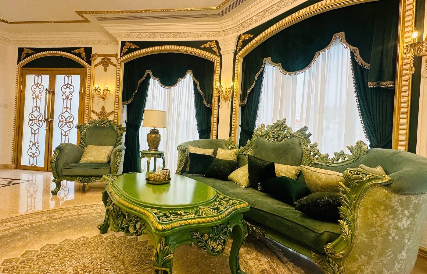 Carved green velvet furniture with gold leaf accents against dark green and sheer white curtains, gold molding and polished marble flooring