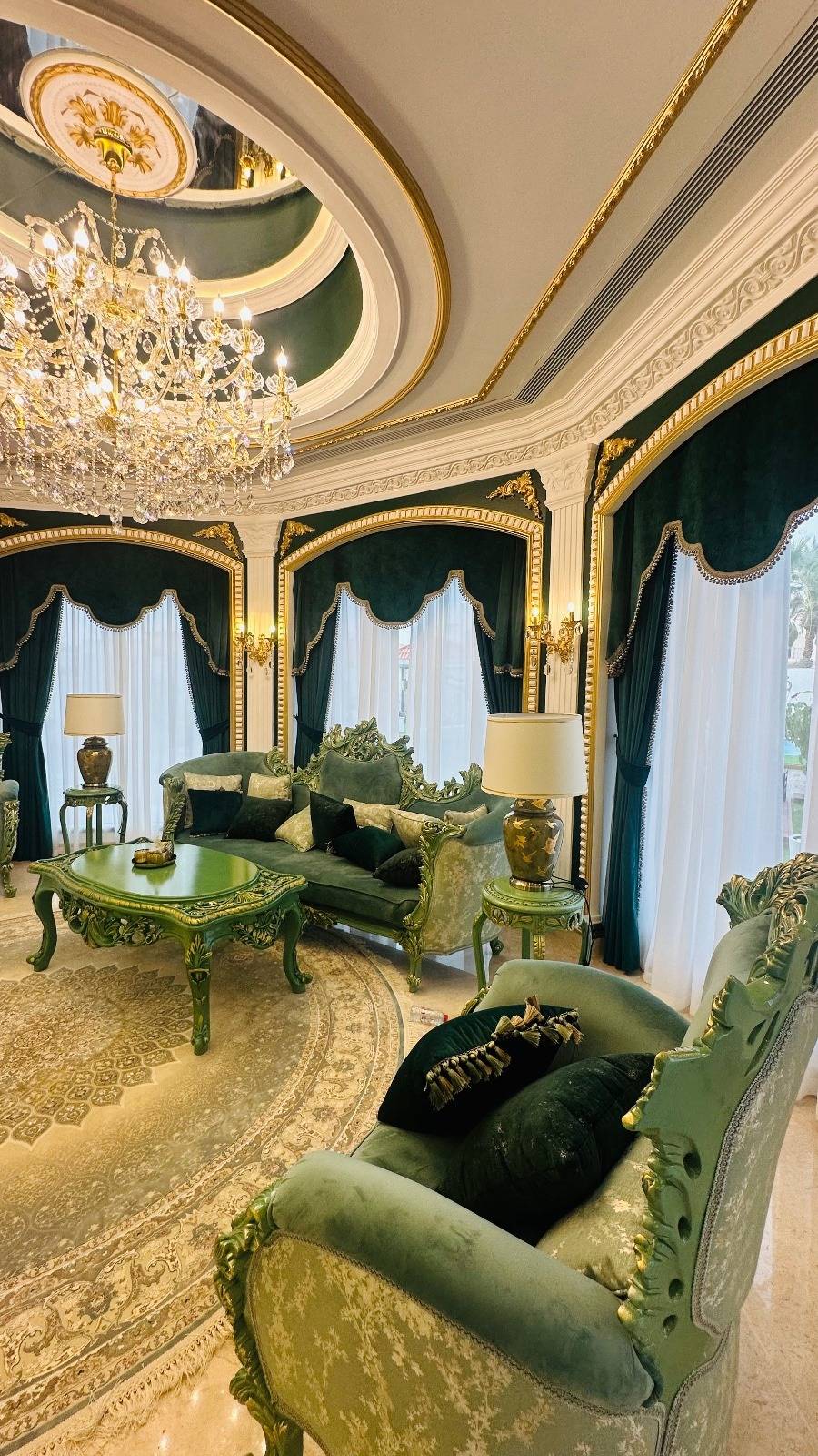 Opulent living room in emerald green and gold with ornate velvet sofas, a matching carved coffee table and a grand crystal chandelier under a decorative ceiling.