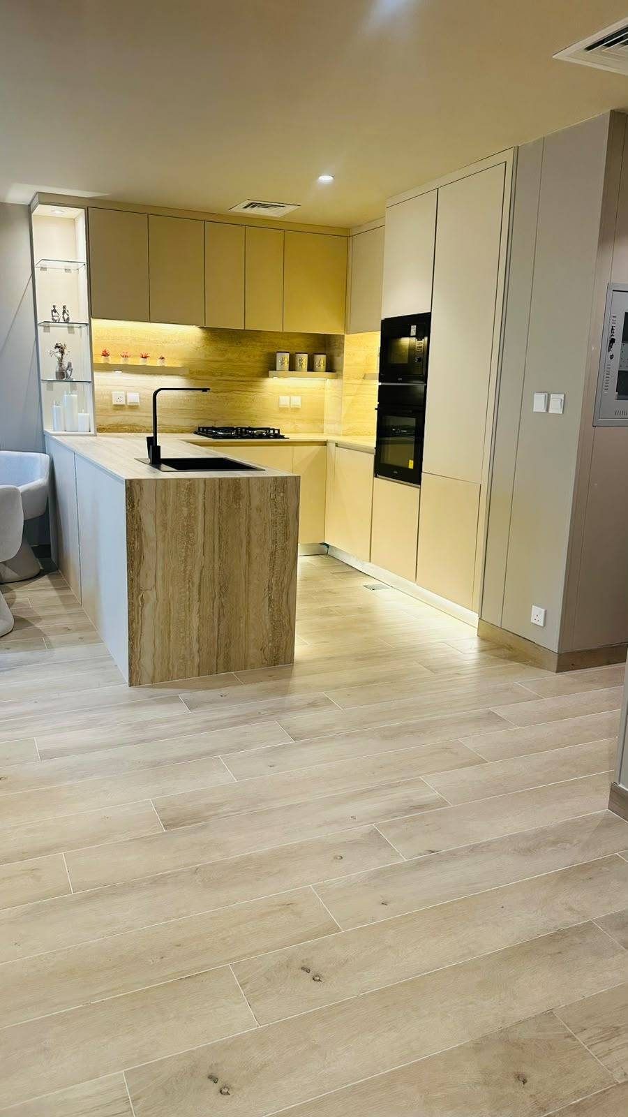 Light beige flat panel cabinetry with a black sink and faucet on a statement island, integrated black appliances and warm under cabinet lighting over a travertine look backsplash