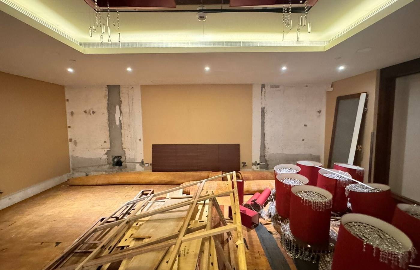 Dubai room mid renovation with stripped concrete walls, a warm ochre accent wall with a dark brown panel, false ceiling lighting and chandeliers, and construction materials in the foreground