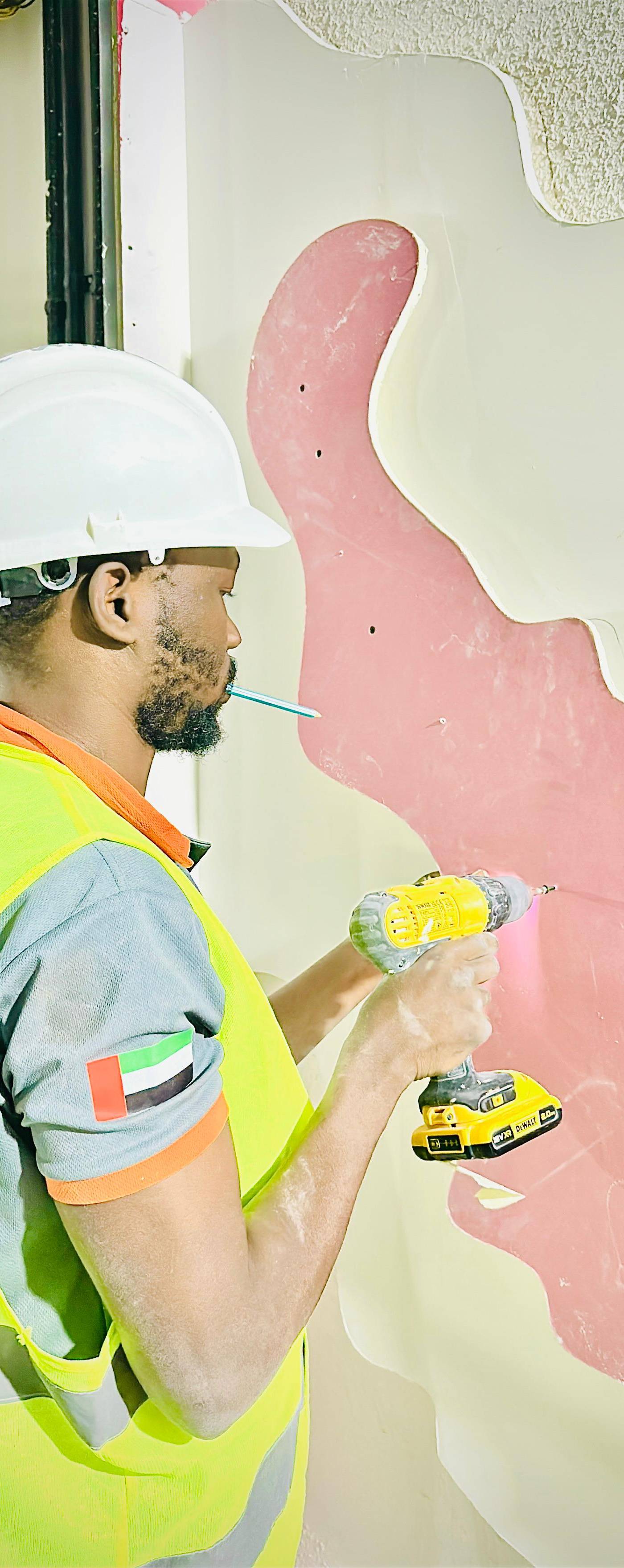 A construction worker wearing a white hard hat and UAE flag high-visibility vest drills into a wall with pink underlayer during an interior fitout.
