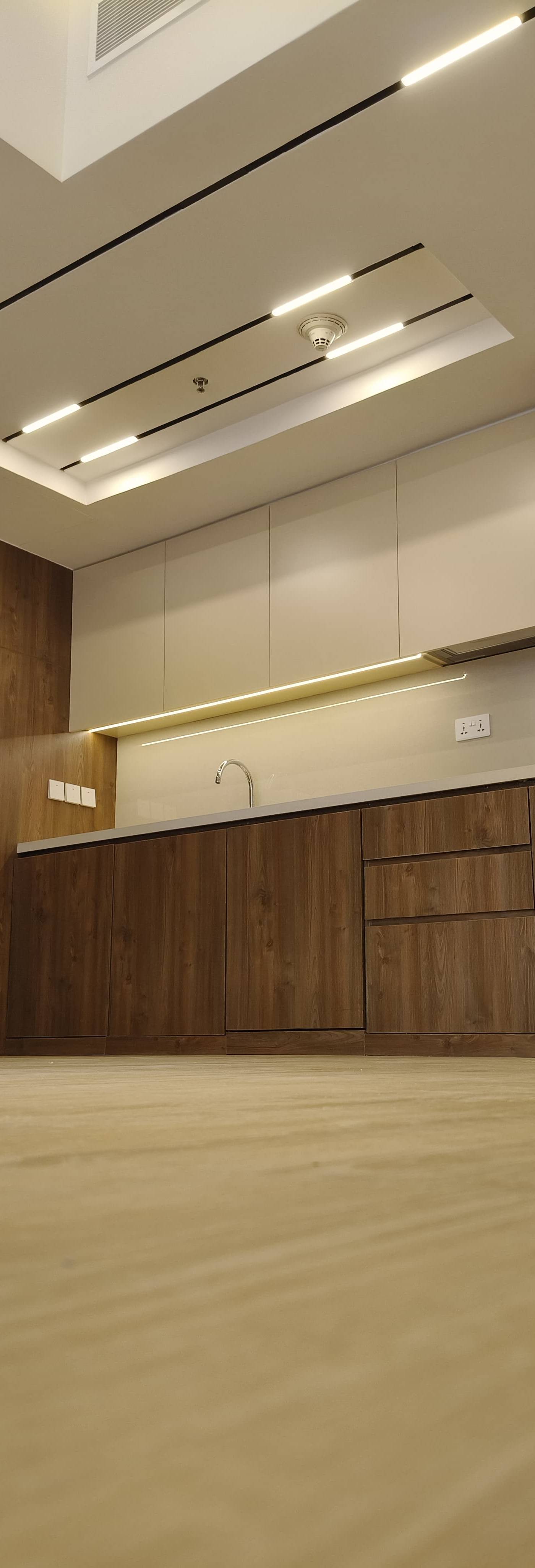 Modern renovated kitchen with warm wood grain lower cabinetry, beige upper cabinets, light grey countertop, integrated oven, and multiple layers of linear LED lighting