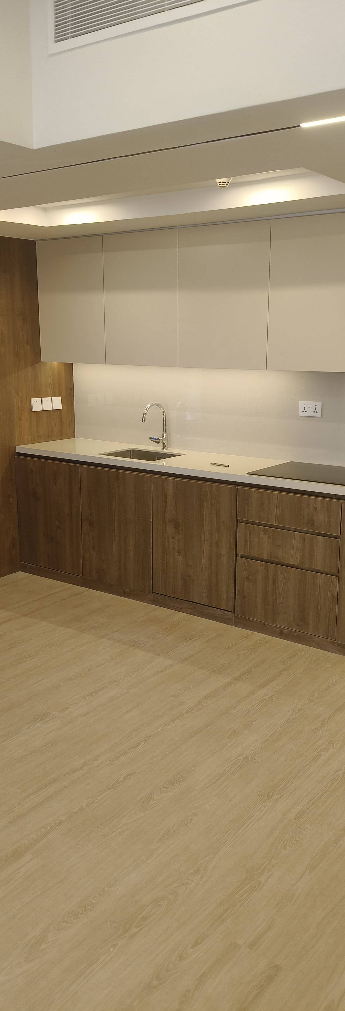 A pristine, freshly renovated modern kitchen features warm wood grain base and tall cabinetry, light beige upper cabinets, a sleek white quartz countertop with an integrated sink and induction cooktop, complemented by a stylish fluted glass pantry cabinet and contemporary linear lighting.