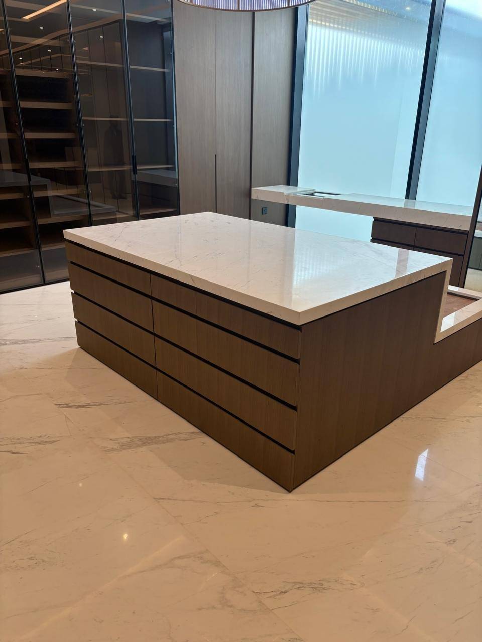 Spacious walk-in closet with central dark wood island, white marble countertop, built-in wardrobes with light wood and tinted glass fronts, and polished marble floor